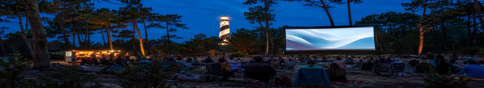 Grand écran de cinéma en plein air face au public, phare landais éclairé en arrière-plan à la nuit tombée